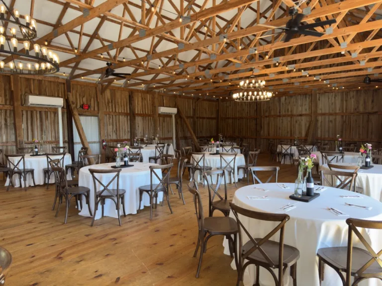 Wide view of the rustic barn reception hall with exposed timber trusses, string lights, and chandeliers above round tables dressed in white linens with dark cross-back chairs and small bottle bud vase centerpieces, set for a corporate 30th anniversary celebration in Martinsburg, WV.