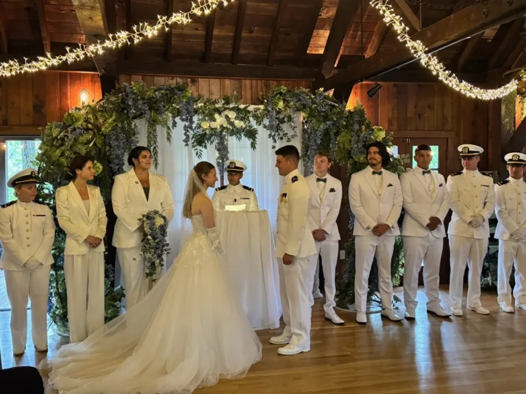 Wedding ceremony with the bride and groom at the altar, the wedding party in white uniforms and a grand floral arch under string lights in Cacapon, WV