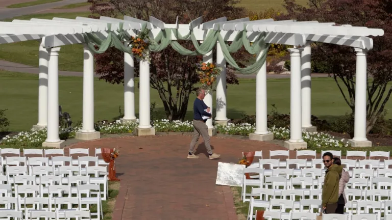 Outdoor wedding ceremony setup with white pergola draped in white fabric and greenery, orange floral arrangements, and rows of white folding chairs on a brick patio