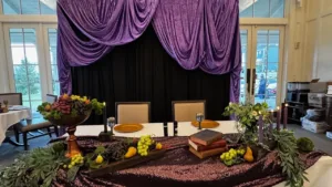 Close-up of head table with fruit compote, stacked vintage books, purple candles and greenery on a sequined runner beneath a purple draped backdrop, 60th birthday party decor in Dumfries, VA