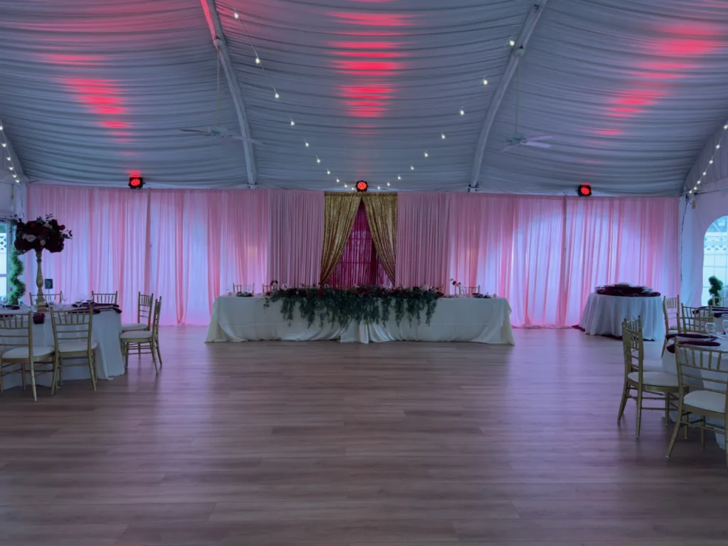 Tented reception hall with draped ceiling, pink uplighting and sheer curtains, head table with greenery and burgundy florals and gold chiavari chairs, quinceañera decor in Columbia, MD