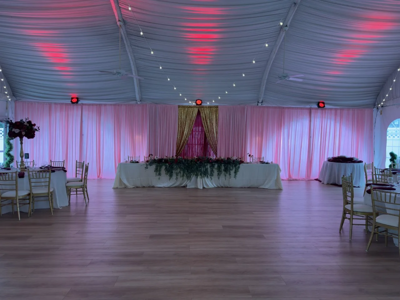 Tented reception hall with draped ceiling, pink uplighting and sheer curtains, head table with greenery and burgundy florals and gold chiavari chairs, quinceañera decor in Columbia, MD