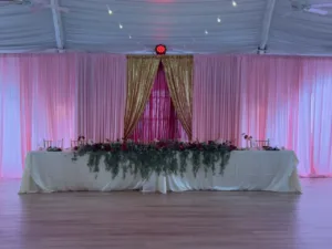 Head table with white cloth, greenery garland and burgundy florals before pink sheer curtains and a gold sequin drape, quinceañera decor in Columbia, MD