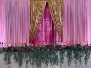 Pink sheer curtains with a gold sequin drape behind a greenery garland with burgundy roses and gold chiavari chairs, quinceañera head table decor in Columbia, MD