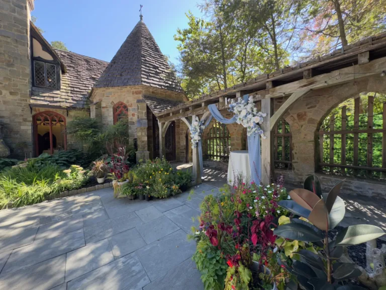 Stone castle tower and pergola with blue draped fabric and white floral arrangements over a white-clothed table amid gardens, wedding decor at The Cloisters Castle in Timonium, MD