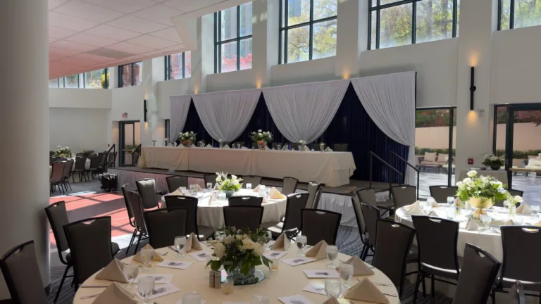 Wide view of quinceanera reception ballroom with round tables in white linens, white floral centerpieces, and head table backed by white and dark drapes