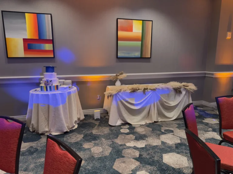 Graduation cake with photo-printed tiers and a blue cap topper on a draped round table, beside a long table with a pampas grass garland and colorful wall art at Mayuri's graduation party in Bethesda, MD