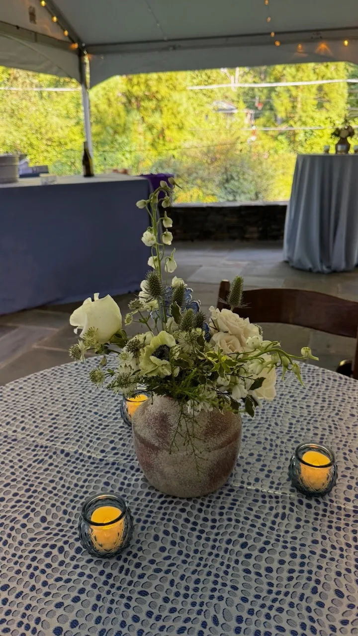 Round table with blue patterned tablecloth, rustic white floral centerpiece in stone vase, and two lit candles under outdoor tent