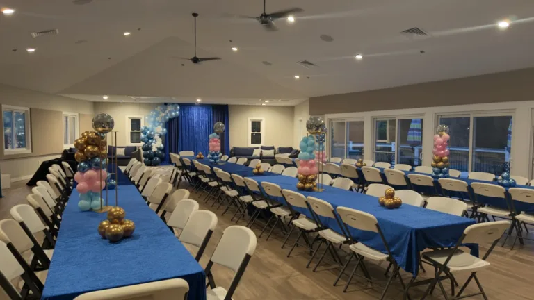 Indoor birthday party room with long tables in blue tablecloths, white folding chairs, and tall blue, pink, and gold balloon centerpieces