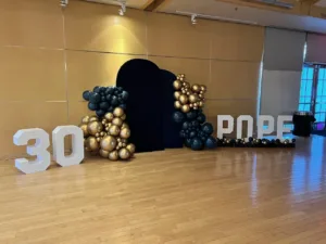 30th birthday decor with white '30' and 'POPE' marquee letters, black and gold balloon garlands around a black arched backdrop, Fairfax VA