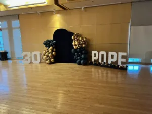 Wide view of 30th birthday party decor with '30' and 'POPE' marquee letters, black and gold balloon columns flanking a black arched backdrop, Fairfax VA