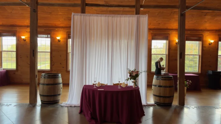 Wedding sweetheart table with burgundy tablecloth in front of white draped fabric backdrop, flanked by wine barrels, in rustic wood venue, Mt. Airy MD