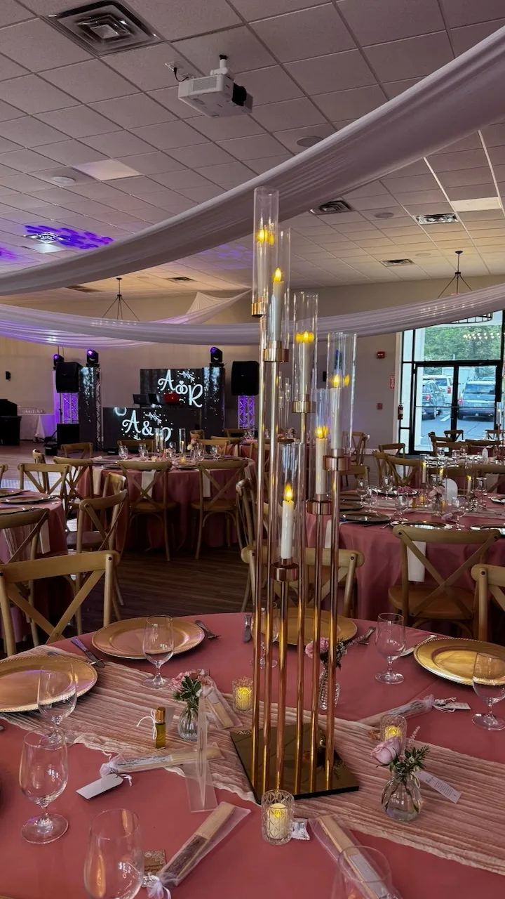 Reception table with pink tablecloth, gold chargers, and tall brass candle holders with lit candles, DJ booth with monogram in background